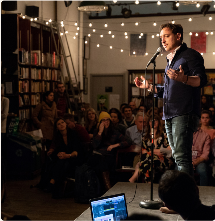 A storyteller speaking at a microphone on a lamplit stage
