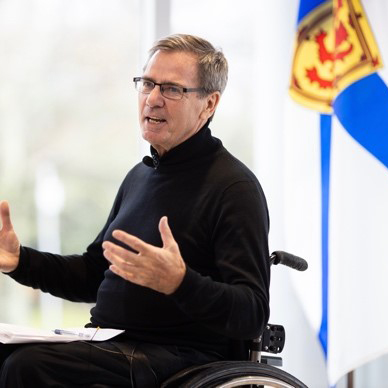 Photo of Gerry Post delivering a presentation in his wheeldchair. A Nova Scotia flag is in the background