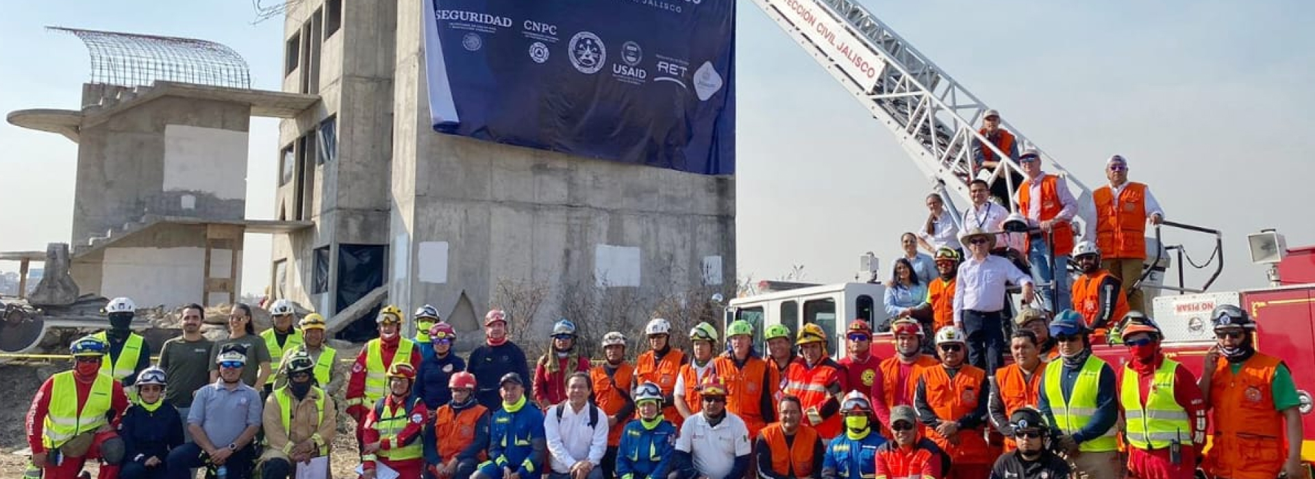 Photo of a range of local, national and regional participants in a search and rescue event in Guadalajara, the rescuers are lined up in front of a fire truck at a disaster simulation site.